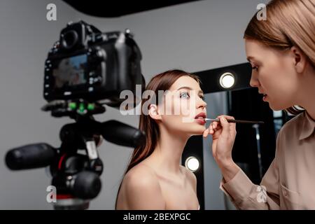 Selektiver Fokus von Make-up-Künstler Anwendung Lipgloss auf Lippen des attraktiven Modell in der Nähe von Digitalkamera auf grau Stockfoto