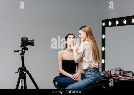 Fröhliche Make-up Künstler Anwendung Lipgloss auf Lippen des attraktiven Modell und Blick auf Digitalkamera auf grau Stockfoto
