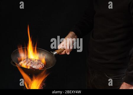 Junger Bursche in Pullover und Jeans. Er hält brennende Wok-Pfanne über Feuer und braten Rindfleisch Steak, vor schwarzem Studio Hintergrund. Seitenansicht Stockfoto
