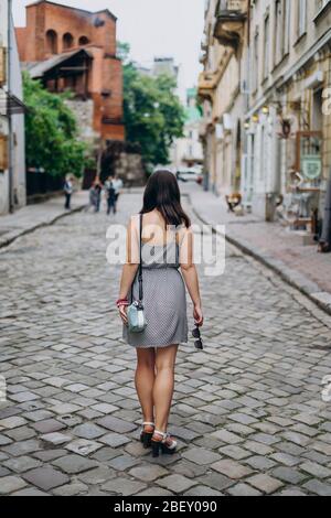 Junge Frau in einem grauen Sonnenkleid auf einer Stadttour. Brunette Frau in Kleid und Sandalen zu Fuß auf Straßen der Stadt. Mädchen in einer Handtasche und Brille Stockfoto