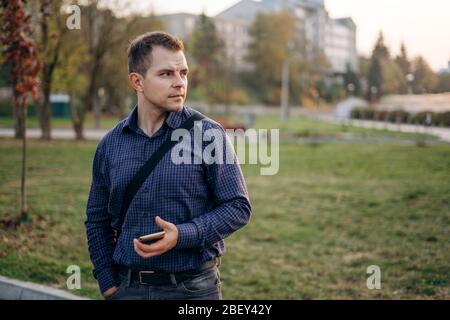 Ein Mann in einem blauen Hemd steht im Park. Ein Mann hält sein Telefon und geht im Park spazieren Stockfoto