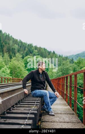 Ein Mann in Jeans sitzt auf einer Bahnstrecke. Eisenbahnstrecke auf Waldhintergrund. Stockfoto