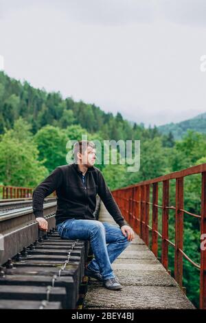 Ein Mann in Jeans sitzt auf einer Bahnstrecke. Eisenbahnstrecke auf Waldhintergrund. Stockfoto