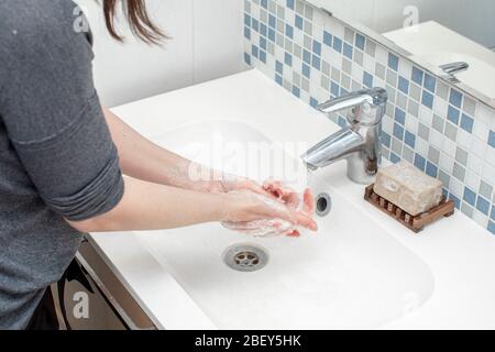 Frau, die ihre Hand mit grüner Seife vor ihrem weißen Waschbecken wäscht. Wasser fließt durch den silbernen Wasserhahn. An der Wand sind kleine Fliesen Stockfoto