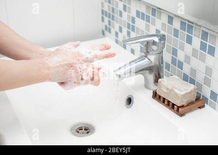 Frau, die ihre Hand mit grüner Seife vor ihrem weißen Waschbecken wäscht. Wasser fließt durch den silbernen Wasserhahn. An der Wand sind kleine Fliesen Stockfoto