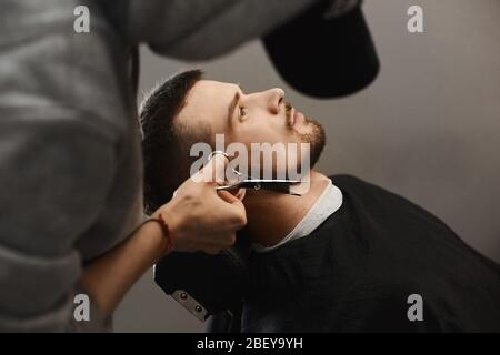 Barbier Mann trimmt stilvollen Bart des schönen jungen brutalen Mann mit modischen Haarschnitt, der im Sessel sitzt bei einem Friseursalon Stockfoto