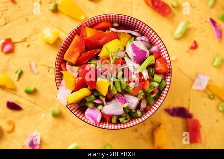 Hochwinkeliger Blick auf eine Schüssel mit einer Mischung verschiedener roher gehackter Gemüse, wie Spargel, Zwiebel oder gelber und roter Paprika, auf goldener Textur Stockfoto