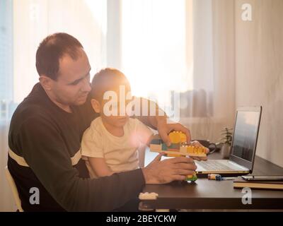 Vier Jahre alter Junge spielen und Vater am Tisch mit Notebook. Home Arbeiten bei Coronavirus Quarantäne Isolationsperiode. Arbeiten unter Kindern, Arbeiten Remote-Konzept. Sonnenuntergang Licht Stockfoto