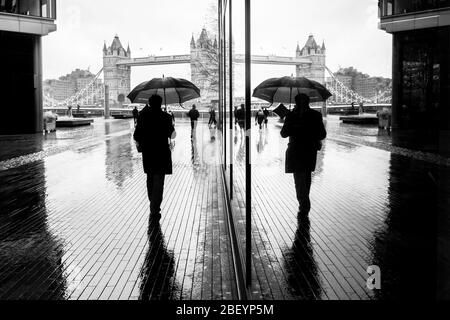 London schwarz-weiß-Straßenfotografie: Mann mit Regenschirm und Tower Bridge Reflexion. Stockfoto