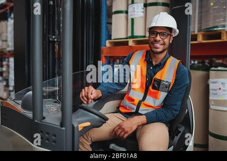 Lächelnder Gabelstaplerfahrer mit Brille im Fahrzeug im Lager, die auf die Kamera schaut, die einen weißen Helm und eine Sicherheitsweste trägt Stockfoto