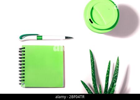 Flacher Einlegeweg mit beschnittenem grünen Notizblock, Stift, wiederverwendbarer Kaffeetasse und Aloe Vera Pflanze auf weißem Hintergrund. Stockfoto