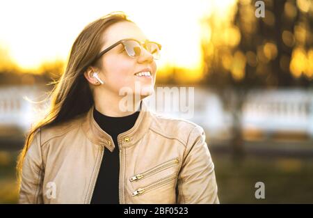 Kabellose Kopfhörer, Ohrhörer. Frau, die in einem Park Musik mit Ohrhörern hört. Glückliche junge Dame lächelnd. Sonnenbrille und Lederjacke. Stockfoto