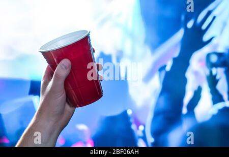 Rote Party-Tasse in der Hand in Nachtclub, Bar oder College-Studenten-Event. Bierkrug aus Kunststoff. Leute, die Spaß in blauen Nachtclub Disco Lichter auf der Tanzfläche. Stockfoto