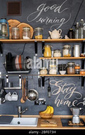 Design der modernen stilvollen Hausküche im rustikalen Loft-Stil und. Schwarze Kreidewand mit Regalen, Tabletts, Gläsern, Tassen, Spüle, Herd und Spüle. Stockfoto