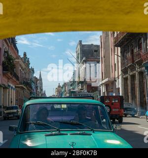 Ein Paar in einem lada Auto, das die Daumen hoch zeigt, wenn Touristen auf dem Rücken eines gelben Cocotaxi durch die Altstadt von Havanna fahren Stockfoto