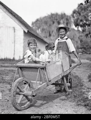 1930ER LÄCHELNDER JUNGE TRÄGT STROH HUT SCHIEBEND BAUERNHOF HOLZRAD BARROW MIT ZWEITEN JUNGEN UND HUND SITZEN IN IHM BLICK AUF DIE KAMERA - D3649 HAR001 HARS BALANCE COMIC TEAMWORK ERFREUT FREUDE LIFESTYLE BRÜDER LÄNDLICHE GESUNDHEIT KOPIEREN RAUM FREUNDSCHAFT VOLLE LÄNGE HALBE LÄNGE OVERALLS PFLEGE LANDWIRTSCHAFT MÄNNER GESCHWISTER SCHUBKARRE VERTRAUEN AUSDRÜCKE LANDWIRTSCHAFT B & W AUGENKONTAKT HUMORVOLLES GLÜCK SÄUGETIERE FRÖHLICHE STÄRKE UND ECKZÄHNE AUFREGUNG AUSSEN BAUERN ERHOLUNG KOMISCH BARROW IN IHM GESCHWISTER LÄCHELT POOCH KONZEPTIONELLE STROH HAT KOMÖDIE FREUDIG ZWEITE STILVOLLE UNTERSTÜTZUNG MUTT WHEEL BARROW EIN WEITERER HUND Stockfoto