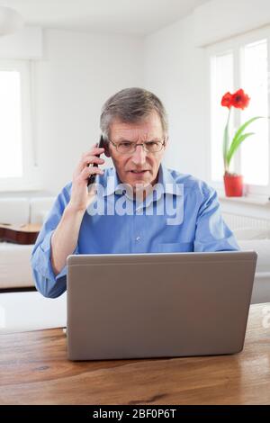 Ernst reifen Geschäftsmann zu Hause mit Laptop arbeiten, während im Gespräch auf dem Handy - konzentrieren Sie sich auf das Gesicht Stockfoto