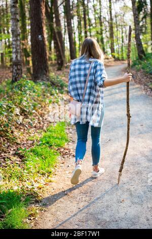 Junge Frau, die durch den Wald wandert. In der Hand hält sie einen Spazierstock. Blick von hinten mit Hintergrundbeleuchtung. Ort: Deutschland, Nordrhein-Westp Stockfoto