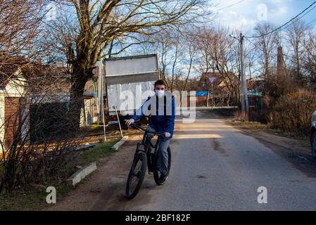 Ein junger Mann in einer schützenden medizinischen Maske und Gummihandschuhen fährt ein Fahrrad entlang einer Dorfstraße. Stockfoto