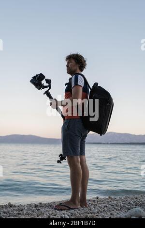 Junger männlicher Fotograf steht am Kiesstrand am Abend und hält seine dslr-Kamera auf einem Gimbal-Stick auf der Suche nach einem perfekten Ort, um ein Video zu schießen. Stockfoto