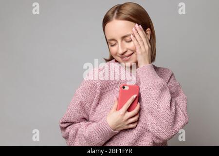 Oh, so süß. Angenehm fröhlich schöne europäische Frau mit Smartphone an Brust und Herz, geschlossene Augen, isoliert auf grauem Hintergrund. Position Stockfoto