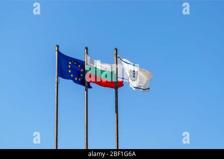 Drei Flaggen auf einem Pfosten gegen hellblauen Himmel: EU, Bulgarien und mit dem Wappen der Stadt Varna an einem sonnigen Tag. Fahnen winken im Wind. Stockfoto