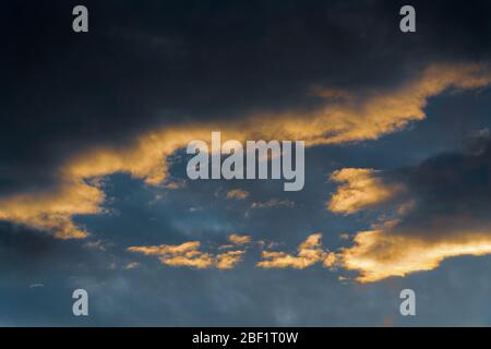 Flauschige Gewitterwolken, die bei Sonnenuntergang von verschwindenden Strahlen beleuchtet werden, und dunkle Gewitterwolken, die über den sonnigen blauen Himmel schweben, um das Wetter der Saison zu ändern Stockfoto