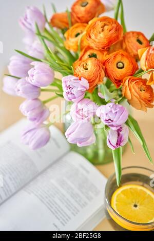 Frühlingskomposition mit lila Tulpen und orangefarbenen Butterblumen und Tasse grünem Tee mit Zitrone, ein offenes Buch auf einem Holztisch. „zu Hause bleiben“-Konzept Stockfoto