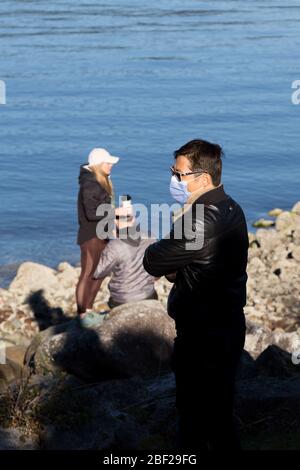 NORTH VANCOUVER, BC, KANADA - APR 11, 2020: Ein Asiat, der während der Pandemie Covid 19 eine medizinische Gesichtsmaske mit einem jungen Paar im Hintergrund trägt Stockfoto