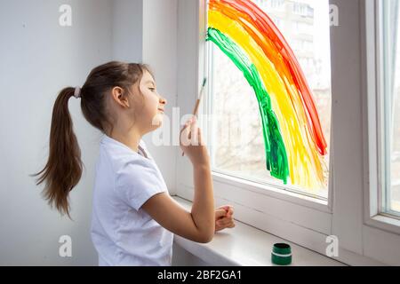 Das Kind zeichnet einen Regenbogen auf das Fenster, um zu zeigen, dass alles gut wird. Jagt den Regenbogen-Fashmob. Quarantäne während eines Pandemiekonzepts. Innen Stockfoto