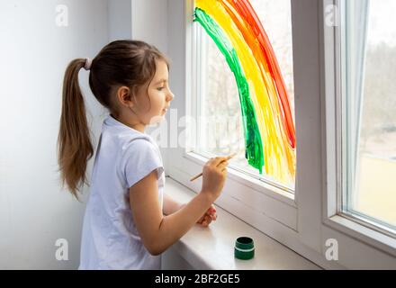 Ein Mädchen mit dunklen Haaren zeichnet einen Regenbogen auf das Fenster. Ein Symbol der Hoffnung, dass alle gesund sein und die Pandemie so schnell wie möglich beenden. Kinder Stockfoto