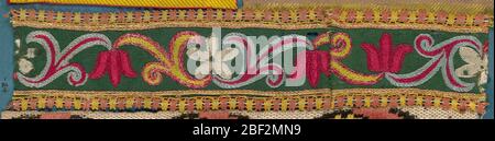 Trimmen. Das Fragment des Schneidens in Form von Tulpen und Blüten mit fünf Blättern, die in einer Kettennaht in rot, gelb, weiß und blau auf grünem Grund gestrickt sind. Schmale Spitzeneinfassung beidseitig in rosa und gelb. Stockfoto