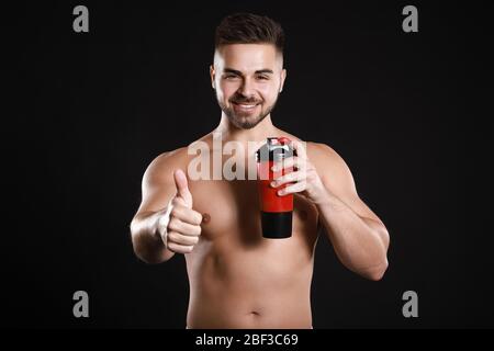 Sportlicher Mann mit Protein-Shake zeigt Daumen-up auf dunklem Hintergrund Stockfoto