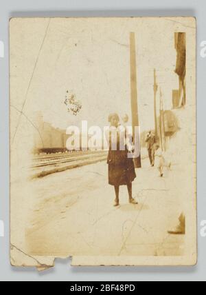 Fotografischer Druck von nicht identifizierter Frau und Kind. Ein Schwarz-Weiß-Foto einer nicht identifizierten Frau, die ein unidentifiziertes Kind hält. Sie stehen auf dem Bürgersteig in der Nähe von Bahngleisen. Ein weiteres nicht identifiziertes Kind und ein nicht identifizierter Mann sind im Hintergrund sichtbar. Stockfoto