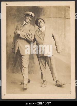 Foto von zwei Männern, die Arme verbinden. Ein Schwarz-Weiß-Foto von zwei Männern, die Arme verbinden.die beiden Männer stehen in der Mitte des Bildes. Beide Männer tragen helle dreiteilige Anzüge mit flachen Kappen. Stockfoto