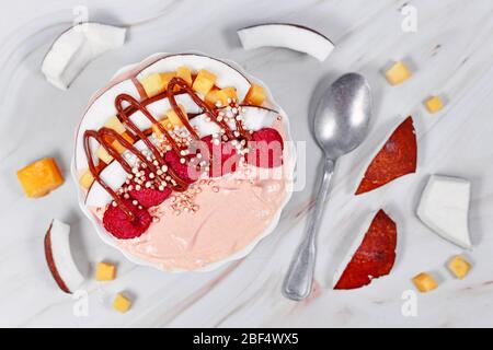 Gesunde Smoothie-Schüssel mit Kokosnussscheiben, Mangostücken und roten Himbeerfrüchten, gekrönt mit gepufften Quinoa-Körnern und Schokoladensauce Stockfoto