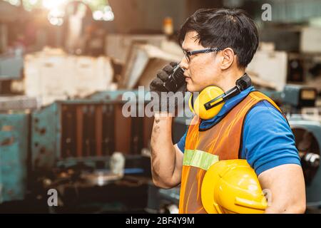 Asiatischer Techniker mit Schutzanzug arbeitet im Werk hart an der Kommunikation mit dem Radio, das Kamera und Lächeln sieht. Stockfoto