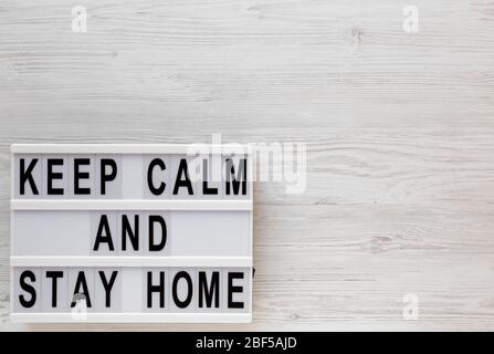 „Keep Calm and Stay Home“-Worte auf einem modernen Brett auf weißem Holzhintergrund, Draufsicht. Über Kopf, von oben, flach gelegt. Kopierbereich. Stockfoto