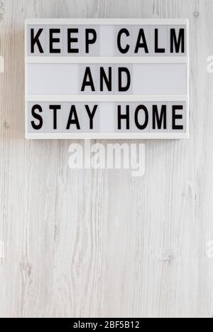 „Keep Calm and Stay Home“-Worte auf einem modernen Brett auf einer weißen Holzfläche, Draufsicht. Über Kopf, von oben, flach gelegt. Kopierbereich. Stockfoto