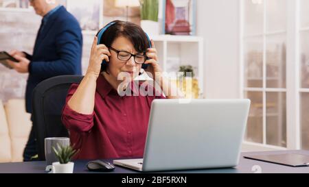 Alte Frau, die eine Tasse Kaffee beim Arbeiten mit einem Laptop mit Kopfhörern auf dem Kopf genießt. Älterer Mann, der im Hintergrund eine Tablette verwendet. Stockfoto