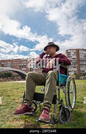 Kaukasischer Mann mit Brille und Hut depressiv und traurig in einem einsamen Rollstuhl in einem Park mit Himmel und einigen Gebäuden im Hintergrund mit Raum Stockfoto