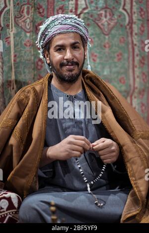 Porträt eines Beduinen-Mannes in traditioneller Kleidung, der mit einem tasbih betet, während er nachts in der saudischen Wüste Tee auf einem Teppich trinkt Stockfoto
