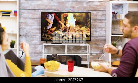 Blick von hinten auf das junge Paar, das sich auf dem Stuhl entspannt und beim Fernsehen beim Essen von Pommes und Popcorn zuschaut. Katze vor dem fernseher. Stockfoto