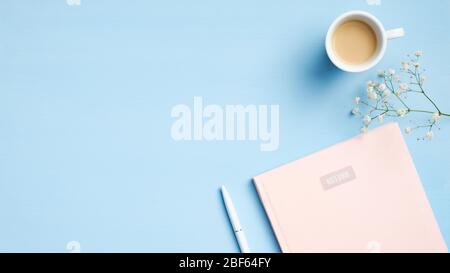 Minimaler femininer Arbeitsplatz mit pinkfarbenem Notizbuch, Tasse Kaffee, Stift, Blumen auf blauem Hintergrund. Moderner Schreibtisch. Draufsicht mit Kopierraum, FLA Stockfoto