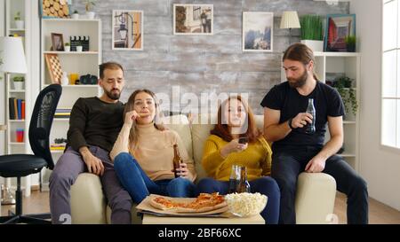 Mann, der nach seinem Bier greift, während er mit seinen Freunden auf der Couch fernsehen sieht. Frauen essen Pizza und Chips. Stockfoto