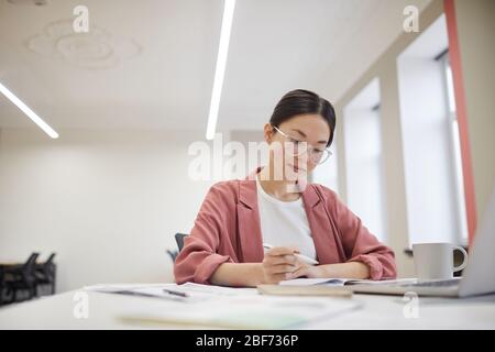 Junge asiatische Geschäftsfrau in Brillen sitzt an ihrem Arbeitsplatz und schriftlich in Dokumenten im Büro Stockfoto