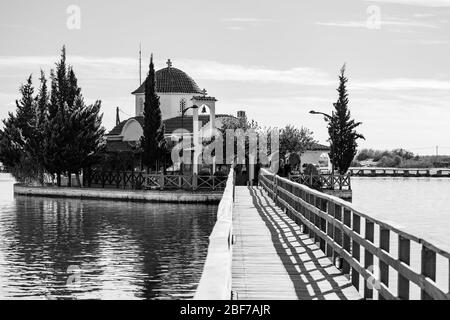 PORTO LAGOS, GRIECHENLAND - 29. NOVEMBER 2019: Touristen genießen schöne Kloster St. Nikolaus am See Vistonida, Porto Lagos, Xanthi Region in Nordgriechenland, schwarz-weiß Blick mit Wasserbrücke Stockfoto