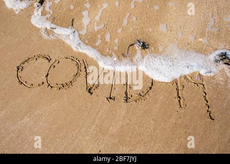 Covid-19 geschrieben in Sand, der von eingehender Flut weggespült wird. Zur Veranschaulichung des Konzepts der Coronavirus-Pandemie. Großbritannien Stockfoto