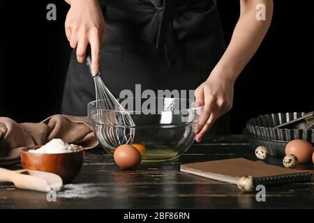 Frau mit Kochbuch macht Teig am Tisch auf dunklem Hintergrund Stockfoto
