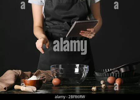 Frau mit Kochbuch macht Teig am Tisch auf dunklem Hintergrund Stockfoto